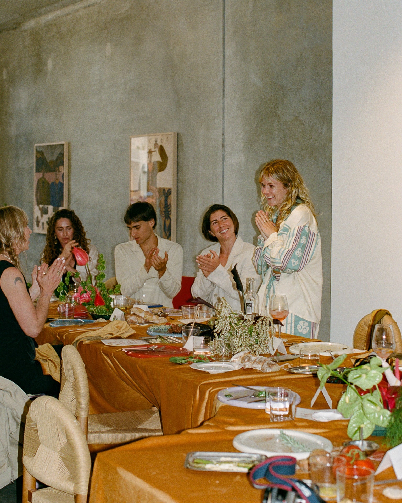 Group of people sitting around a dining table with a neutral wall and framed pictures in the background.