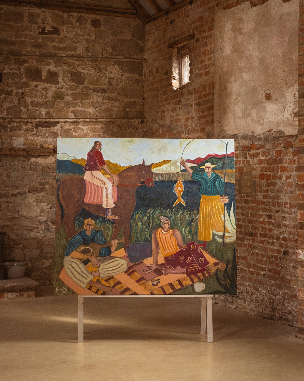 A large colorful painting depicting figures, animals, and landscapes, displayed on a white stand inside a rustic stone and brick gallery room.